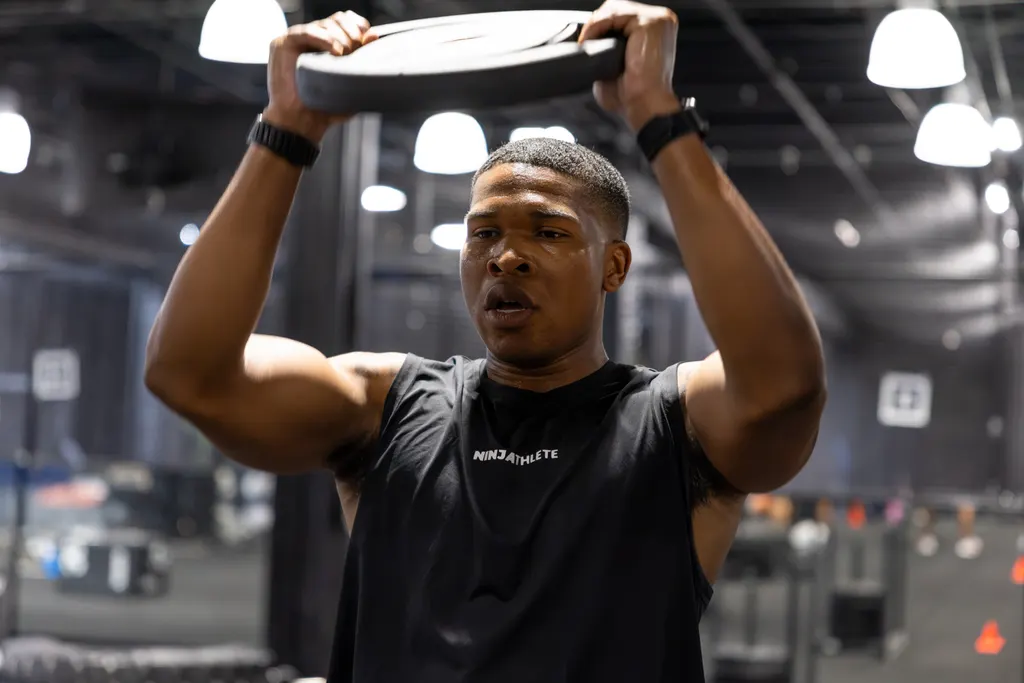 Athlete pressing a weight plate overhead on the training floor at Dallas Performance Systems